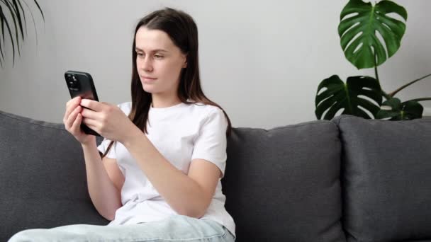 使用智能手机浏览社交媒体 查询新闻 坐在沙发上玩手机游戏等方式来放松快乐的年轻女性 笑一个20多岁的高加索女孩 用手机技术呆在家里 — 图库视频影像