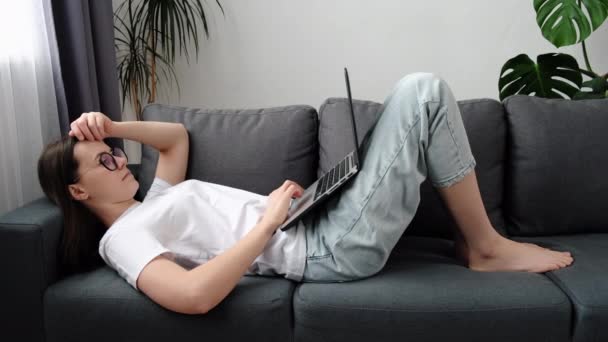 松懈的年轻高加索女人戴着眼镜躺在灰色沙发上 带着笔记本电脑 在社交媒体网络上在线聊天 发电子邮件 懒洋洋地一个人在家里度过周末 互联网使用概念 — 图库视频影像