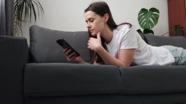 Mutlu gülümseyen, rahat, akıllı telefon kullanan, cep telefonu kullanan, komik videolar izleyen, gri koltukta uzanan, tembel, genç bir kadın, sosyal medyada sohbet eden, evdeki rahat koltukta dinlenen,