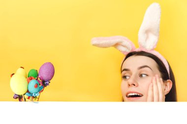 Oldukça etkilenmiş beyaz bir kadının yakın portresi tavşan tüylü kulaklar takıyor, Paskalya renkli yumurtalara bakarken şok edici bir şekilde eğleniyor, stüdyoda sarı renkli arka planda fotokopi ile izole edilmiş pozlar veriyor.