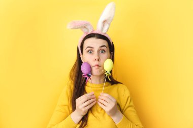 Mutlu, güzel, gülümseyen, 20 'li yaşlarda tavşan kulağı takan, renkli Paskalya yumurtaları tutan, tavşan kulaklı, esmer, sarı arka plan duvarında izole edilmiş poz veren genç bir kadın.