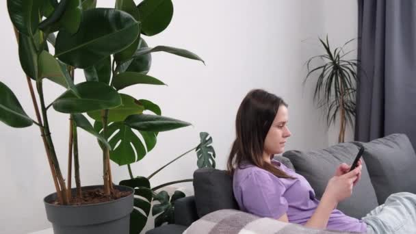 穏やかな若い女性の側面ビューは スマートフォンを保持し リビングルームでソファに座っている千年紀の女性は ソーシャルメディアネットワークを使用してチャット 現代の無線技術と楽しんでインターネット上の商品を選択します — ストック動画