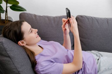 Modern teknoloji aşırı kullanımı. Rahat, genç ve güzel bir bayan cep telefonuyla kanepede uzanıyor. Oturma odasında dinleniyor, kablosuz cihaz kullanıyor, internetten bilgi indiriyor. Harika bir uygulama öğreniyor.