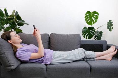 Oturma odasında kanepede uzanmış, akıllı telefon tutan, sohbet eden, sosyal medya ağını kullanan, internetten mal seçen, modern kablosuz teknolojiyle eğlenen genç bir kadının yan görüntüsü.