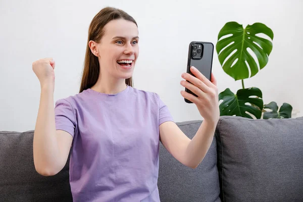 Girl winning on line lottery, received results online auction. Overjoyed young female sit on couch at home holds smartphone reading unbelievable news in sms, celebrate moment of victory feels excited