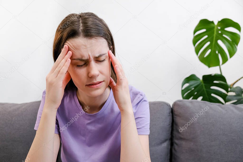 Unhappy tired young caucasian female 20s sitting on couch rubs temple, closed eyes suffers headache painful feelings chronic migraine hurt. Break up and divorce, difficult period of life concept