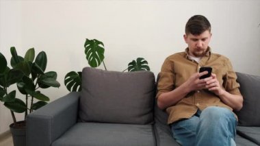 Evdeki gri koltukta oturan genç adam elinde akıllı telefon tutarak SMS 'den kötü haberler aldığını düşünüyor. Aygıt arızası nedeniyle olumsuz duygular, hata bilgisi arızası, kayıp internet bağlantısı kavramı