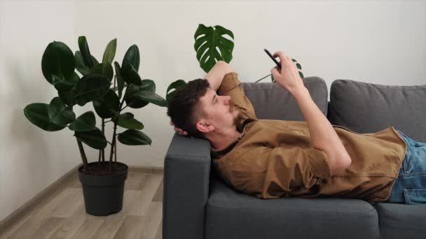 沉着的男人休息用手机查看网上的电子邮件阅读新闻 松懈的年轻人躺在客厅柔软舒适的沙发上 懒洋洋地周末在手机屏幕上看视频 — 图库视频影像