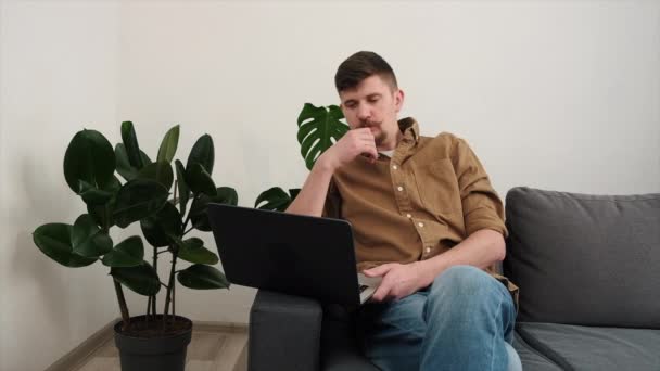 认真的年轻学生自由职业者舒适地坐在家里的沙发上 带着笔记本电脑 在线远程办公的概念 以男性企业家为重点的远程工作 在计算机上打字 领导遥远的通信 — 图库视频影像