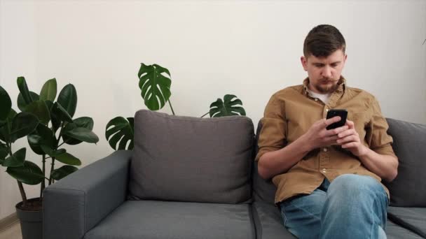 ホームホールドスマートフォンのグレーのソファに座っている若い男は Smsによって恐ろしいニュースを受け取ることについて感じています 負の感情が原因デバイスが破損し エラー情報が失敗し インターネット接続の概念を失った — ストック動画