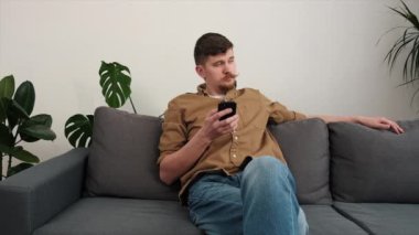 Sakin bir genç adam evdeki oturma odasında rahat bir kanepede dinleniyor, mobil uygulama kullanıyor. Sosyal ağda sohbet eden yakışıklı beyaz bir adam. Erkek arkadaş bulma sitesi ya da internet sörfü konsepti