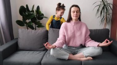 Stressiz konsept. Genç anne evde rahat bir kanepeye oturup Lotus pozisyonunda meditasyon yapıyor, küçük kız yanında zıplayarak eğleniyor. Konsantre anne yogası. Kanepede rahatla. Şakacı çocuğu görmezden gel.