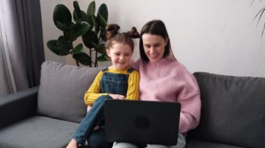 Gülümseyen anne ve küçük kız bilgisayar uygulaması üzerinden webcam ile sohbet ediyorlar. Küçük kızı olan sevimli bir anne el sallıyor uzaktan video çekiyor dizüstü bilgisayarına bakıyor kanepeye oturuyor.