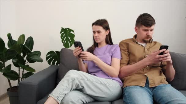 年轻夫妇手持智能手机 看手机 玩手机游戏 一起坐在客厅舒适的灰色沙发上 押注或竞拍拍卖申请表 — 图库视频影像