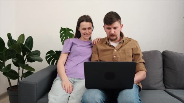 快乐的丈夫和妻子在舒适的沙发上悠闲自在地坐在家里 享受着使用笔记本电脑的乐趣 可爱的年轻夫妇在电脑屏幕上浏览互联网 选择一起上网购物 — 图库视频影像