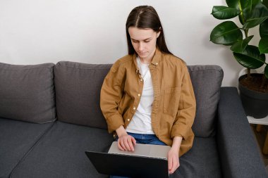 Düşünceli genç bir kadın rahat bir koltukta oturup dizüstü bilgisayarda çalışıyor, evde sorunları çözmeyi düşünüyor, ciddi beyaz kadın 20 'li yaşlarda ilham arıyor.