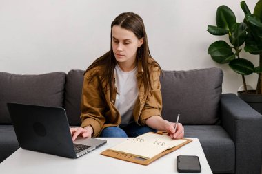 Sanal kurslarda okuyan, uzaktan eğitim alan esmer kız dizüstü bilgisayara bakıp evdeki rahat koltukta oturmuş Webinar dersini izliyor.