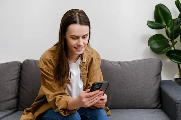Mujer Joven Sonriente Relajarse Sentarse Sofá Casa Usando Teléfono Celular —  Fotos de Stock