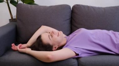 Mutlu pozitif genç bayan rahat bir kanepede düştü, tembelce hafta sonu istirahatinin tadını çıkarıyor evde yalnız başına. Oturma odasındaki rahat gri kanepede dinlenen gülümseyen tatlı kadına yakın durun. Konsept yeniden yükleniyor