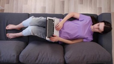 Yukarıdan bakıldığında, koltukta dizüstü bilgisayarı olan gevşemiş genç bir kadın. Milenyum kadınları haftasonunu internet Wi-Fi bağlantısı ve modern kablosuz bilgisayar, sosyal medyada sohbet ve web sörfü kavramını kullanarak geçiriyor