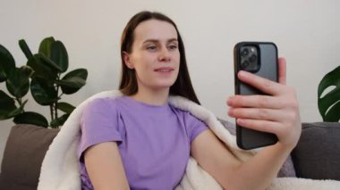 Mutlu genç kadın vlogger elinde akıllı telefon kamerayla video bloğu çekiyor, evdeki rahat koltukta oturmuş görüntülü konuşma uygulaması ile sohbet ediyor.
