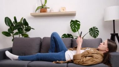 Sakin genç kadın 20 'li yaşlarda rahat bir kanepede uzanıyor modern akıllı telefon kullanıyor, esmer kız rahat gri koltukta dinleniyor cep telefonundan mesaj atıyor ya da sohbet ediyor, cep telefonundan alışveriş yapıyor.