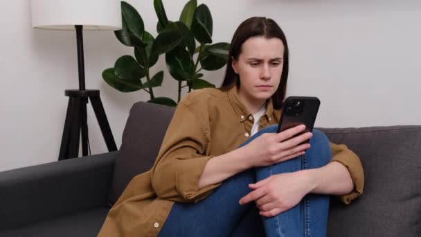 悲しい女の子は スマートフォン上のモバイルメッセージで悪いニュースを読んで動揺している感じ ソファーに座っている不満若い女性は 携帯電話が携帯電話を介していじめられてサイバーいじめに苦しむ恐れを保持しています — ストック動画