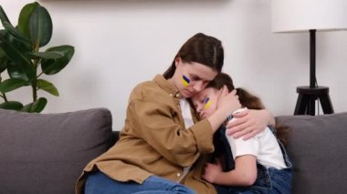 Ukraynalı genç bir annenin Ukrayna bayrağıyla küçük üzgün kızını kucaklaması, evdeki kanepede oturan aile, yoksulluk kavramı. Kriz, barış, saldırganlığı durdurun, Rus savaşına karşı çocuk