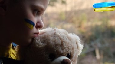 Savaşa karşı çocuk. Yüzünde mavi-sarı bir bayrak olan mutsuz küçük bir kız, elinde tüylü bir ayıyla evde pencere eşiğinde oturan bir çocuk. Sivillerin tahliyesi. Ukrayna 'ya özgürlük. Rus saldırısını durdurun.