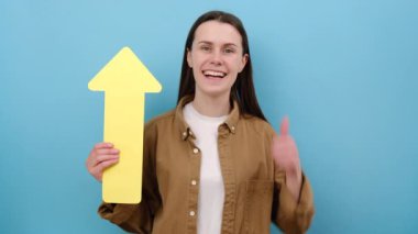 20 'li yaşlarda elinde sarı okla kameraya gülümseyen, büyüyen ve büyüyen bir konseptle bakan, gömlek giyen, stüdyodaki mavi arkaplan duvarının üzerine izole edilmiş pozlar veren genç bir kadın.