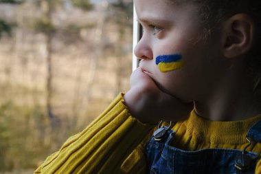 Yüzünde Ukrayna bayrağı olan üzgün küçük kız çocuğunun pencerenin kenarında tek başına oturması. Doğada mutsuz bir çocuk. Barış, savaş yok, yardım edin, Rus saldırısını durdurun. Kriz kavramı