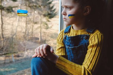 Ukrayna jeopolitik dünya krizi. Mutsuz küçük kız evde pencere eşiğinde tek başına oturuyor. Üzüntü, umut özlemi. Çocuğun yanağında Ukrayna bayrağı var. Savaşı durdurun. Endişe ve beklenti