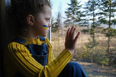 Yüzünde Ukrayna bayrağı olan üzgün küçük bir kız çocuğu. Mutsuz, yorgun bir çocuk. Evde pencere eşiğinde otururken doğa arka planında. Barış, savaş yok, yardım edin, Rus saldırısını durdurun. Kriz kavramı