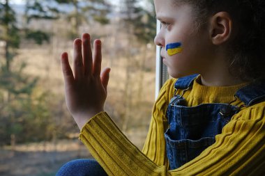 Savaş kavramını durdur. Ukrayna için dua et. Ukraynalı korkmuş çocuk evdeki pencere eşiğinde oturuyor. Üzgün küçük kız patlamalardan korkuyor. Yüzünde Ukrayna bayrağı olan zavallı çocuk üzüntü ve keder hissediyordu.