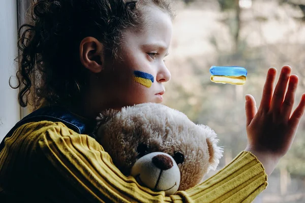 Yüzünde Ukrayna bayrağı olan üzgün küçük bir kız çocuğu, evinde tek başına oturan pofuduk ayıyı tutan yorgun bir çocuk. Barış, savaş yok, yardım edin, Rus saldırısını durdurun. Kriz kavramı