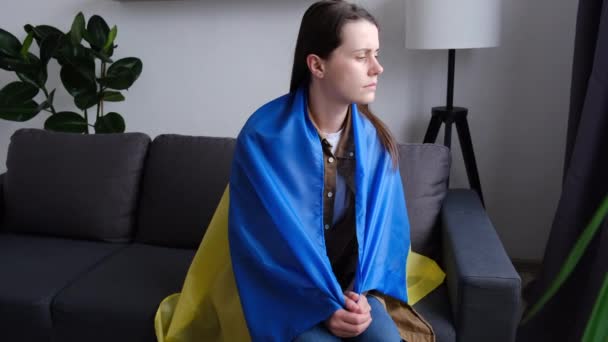 家庭で灰色のソファに一人で座ってウクライナのフラグで覆われた悲しい若い白人女性を動揺 ロシアとの戦争でウクライナ国民と一緒に立っての概念 不安と期待 世界への希望 — ストック動画