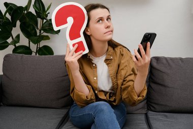 Üzerinde kırmızı soru işareti ve akıllı telefon olan gri koltukta oturan 20 'li yaşlarda, düşünceli, genç beyaz kadın. Çevrimiçi öğrenim, alışveriş, İnternet pazarlama, tefekkür kavramı