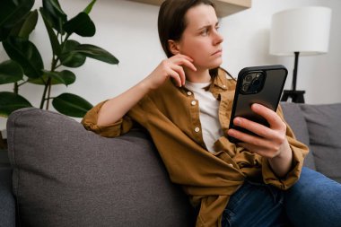 Düşünceli genç bir kadının koltukta oturup, akıllı telefonuna yan gözle bakması, sevgilisinden mesaj beklemesi, yalnız ya da kıskanç hissetmesi. Sosyal medyada haberleri okuyan mutsuz kız