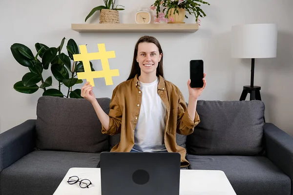 Smiling attractive young woman blogger sitting on comfortable sofa at home showing big yellow hashtag symbol and smartphone, happy looking at camera, sharing viral content, tagged message concept