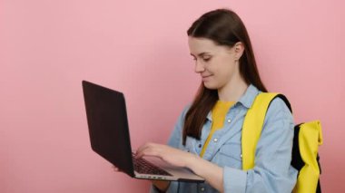 Güzel kız öğrencilerin portresi tişört giyiyor ve sarı sırt çantası bilgisayarında kullanılıyor, baş parmağı görünüyor, pembe arka plan stüdyosu izole edilmiş. Üniversite konsepti eğitimi