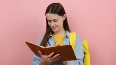 Alıştırma kitabı notlarına yazan çekici genç kız portresi, pembe renkli arka plan stüdyosunda izole edilmiş gömlek ve sarı sırt çantası giyiyor. Üniversite konsepti eğitimi