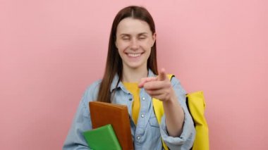 Gülümseyen güzel bir genç kız elinde kitaplarla başparmağını kaldırır başparmağını kaldırır, gömlek giyer ve sarı sırt çantası takar, pembe arka plan stüdyosunda izole bir şekilde poz verir. Üniversite üniversitesinde eğitim