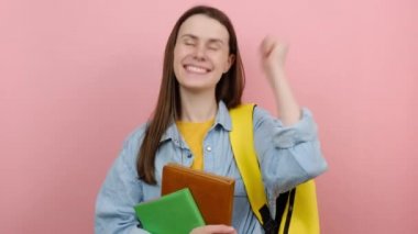 Neşeli genç kız 20 'li yaşlarda tişört giyiyor ve sarı sırt çantalarını tutuyor yumruklarını sıkıyor, soluk pembe arka plan stüdyosuna izole edilmiş poz veriyor. Lise üniversite konsepti eğitimi
