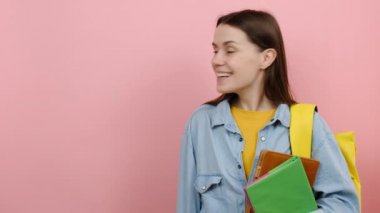 Promosyoncu genç kız 20 'li yaşlarda tişört giyiyor ve sarı sırt çantalı kitaplar çalışma alanını gösteriyor. Fotokopi alanı oluşturuluyor. Stüdyodaki pastel pembe arka plan duvarı üzerinden izole edilmiş poz veriliyor.