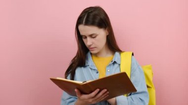 20 'li yaşlardaki kız öğrencilerin portresi gömlek giyer, sarı sırt çantasından kitap okur, hayal kurar, stüdyodaki pembe renkli arka plan duvarında izole bir şekilde poz verir. Lise üniversite konsepti eğitimi