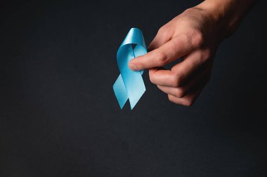 Men's health and Prostate cancer awareness campaign concept. Male hand holding blue ribbon awareness on black background wall in studio with copy space. Symbol for support men who living cancer