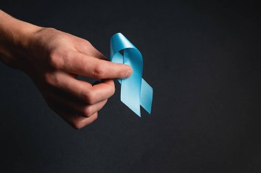 Koyu gri arka plan duvarında mavi kurdeleyi tutan erkek elini kapat. Prostat kanseri farkındalığı, Movember Men 'in sağlık farkındalığı. Sağlık, Uluslararası erkekler, Baba ve Dünya Kanser Günü konsepti