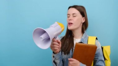 Portrait of promoter girl teen student 20s wears denim jacket and yellow backpack hold books scream in megaphone announces discounts sale Hurry up, posing isolated over blue color background in studio