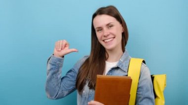 Neşeli, genç kız kot ceket giyiyor ve sarı sırt çantası kitapları tutuyor. Parmağını kendine doğrultuyor. Mavi stüdyo arka planında izole edilmiş pozlar veriyor. Üniversite konsepti eğitimi