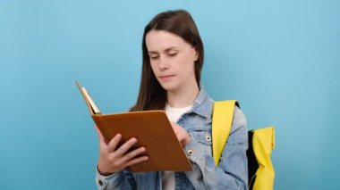 Portre kızı genç kız kot ceket giyiyor ve sarı sırt çantasından kitap okuyor hayal kuruyor, stüdyoda mavi renkli arka plan duvarının üzerinde izole bir şekilde poz veriyor. Lise üniversite konsepti eğitimi